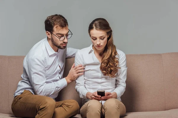 Serious young woman using smartphone while handsome man in eyeglasses sitting on sofa isolated on grey — Stock Photo