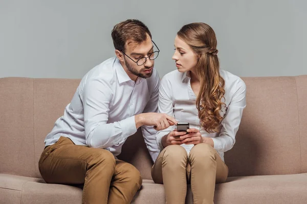 Jovem mulher segurando smartphone e homem bonito em óculos apontando com o dedo enquanto sentado no sofá isolado no cinza — Fotografia de Stock