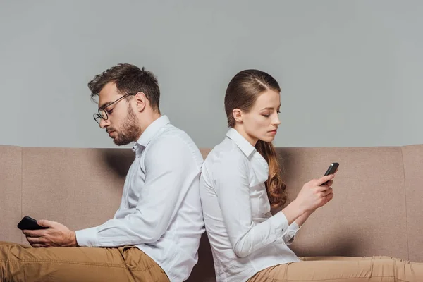 Joven pareja sentado espalda con espalda y el uso de teléfonos inteligentes aislados en gris - foto de stock