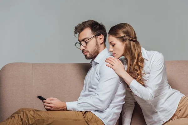 Junge Frau sieht Freund mit Smartphone auf Sofa isoliert auf grau — Stockfoto