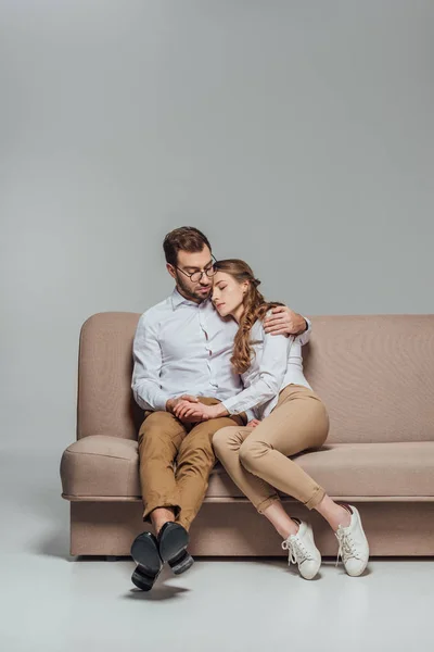 Guapo joven abrazando hermosa novia durmiendo en su hombro mientras está sentado en sofá aislado en gris - foto de stock