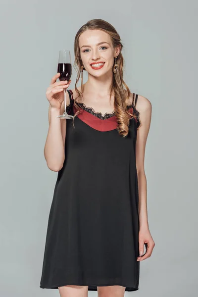 Beautiful young woman holding glass of red wine and smiling at camera isolated on grey — Stock Photo