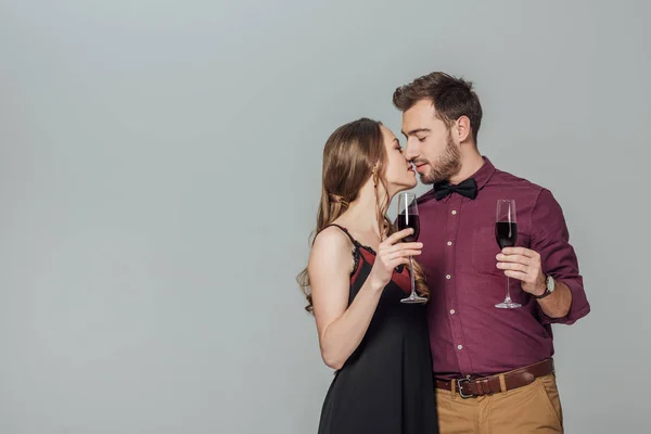 Hermosa pareja joven sosteniendo vasos de vino tinto y besos aislados en gris - foto de stock