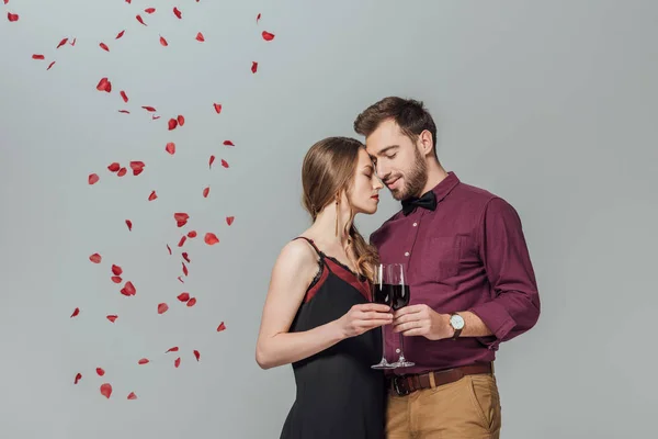 Beautiful young couple holding glasses of red wine and rose petals falling isolated on grey — Stock Photo