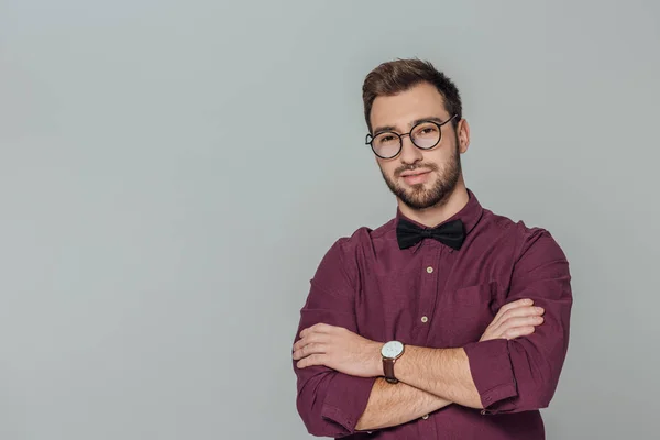 Élégant jeune homme aux lunettes debout avec les bras croisés et souriant à la caméra isolée sur gris — Photo de stock