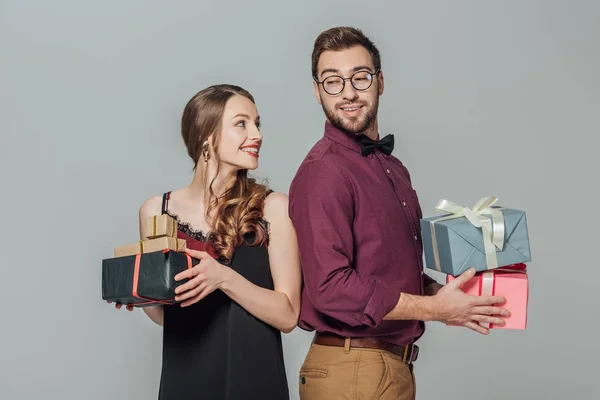 Glückliches stylisches junges Paar hält Geschenkboxen in der Hand und lächelt sich isoliert auf grau an — Stockfoto