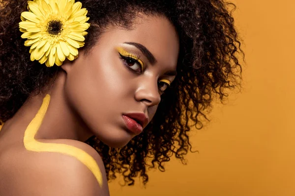 Joven mujer afroamericana tierna con maquillaje artístico y gerbera en el pelo mirando a la cámara aislada sobre fondo naranja - foto de stock