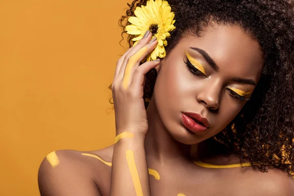 Belle femme afro-américaine avec maquillage artistique et gerbera dans les cheveux rêvant isolé sur fond orange — Photo de stock