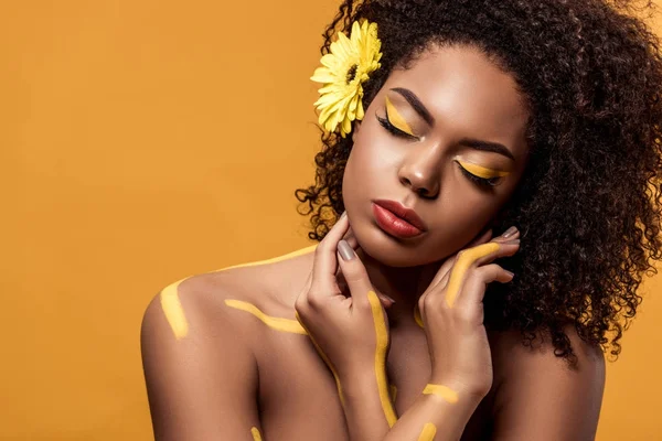 Joven mujer afroamericana sensual con maquillaje artístico y gerbera en el cabello soñando aislado sobre fondo naranja - foto de stock
