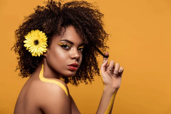 Junge helle afrikanisch-amerikanische Frau mit artistischem Make-up und Gerbera im Haar, die ihr Haar isoliert auf orangefarbenem Hintergrund berührt — Stock Photo