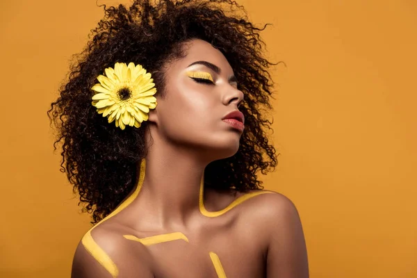 Jovem mulher afro-americana atraente com maquiagem artística e gerbera no cabelo isolado em fundo laranja — Fotografia de Stock