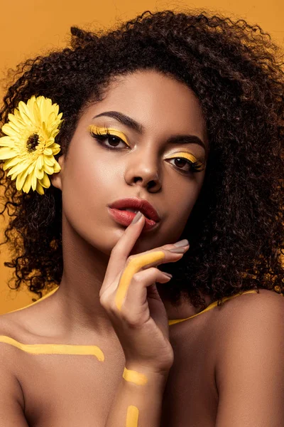 Joven mujer afroamericana sensual con maquillaje artístico y gerbera en el pelo sosteniendo el dedo en los labios aislados sobre fondo naranja - foto de stock