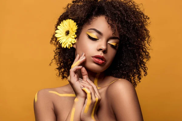 Elegante mujer afroamericana con maquillaje artístico y gerbera en el pelo sostiene las manos cerca de la cara aislada sobre fondo naranja - foto de stock