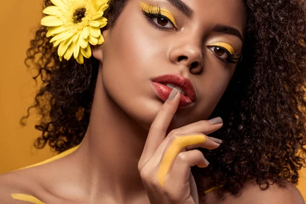 Joven mujer afroamericana sensual con maquillaje artístico y gerbera en pelo con el dedo en los labios aislados sobre fondo naranja - foto de stock