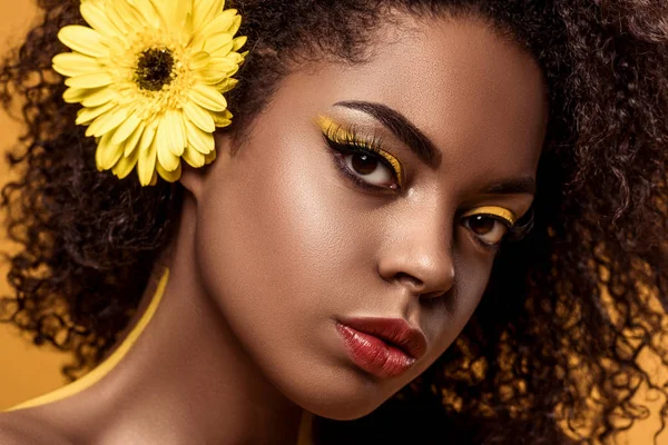 Retrato de cerca de una joven afroamericana sensual con maquillaje artístico y gerberas en pelo aislado sobre fondo naranja - foto de stock