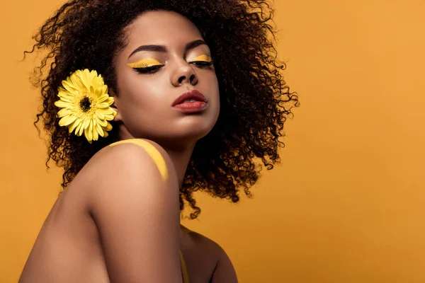 Jeune femme afro-américaine tendre avec maquillage artistique et gerbera dans les cheveux isolés sur fond orange — Photo de stock