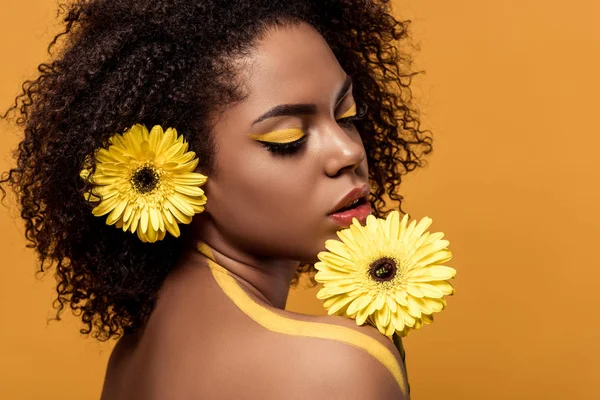 Attractive young american woman with artistic make-up smelling gerbera isolated on orange background — Stock Photo