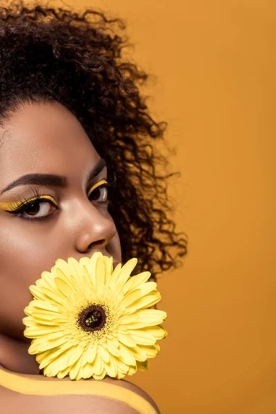 Atraente jovem afro-americana com maquiagem artística mantém flor de gerbera amarela isolada no fundo laranja — Fotografia de Stock