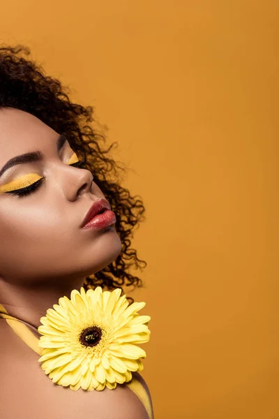 Joven mujer afroamericana brillante con maquillaje artístico sostiene flor de gerberas amarillas aisladas sobre fondo naranja - foto de stock