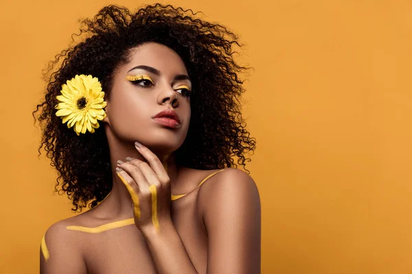 Giovane donna afroamericana sensuale con trucco artistico e gerbera in capelli si tiene per mano dal suo viso isolato su sfondo arancione — Foto stock