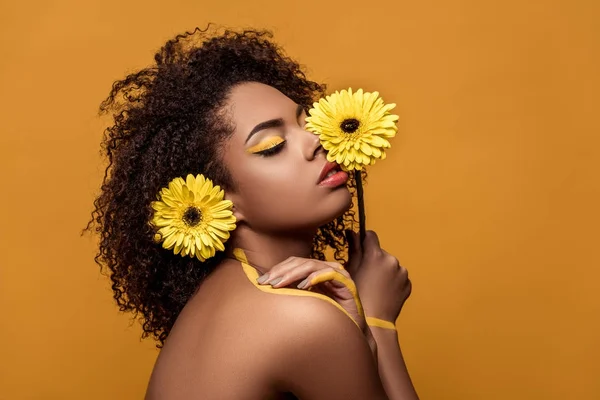 Femme afro-américaine élégante avec maquillage artistique tient fleur par son visage touche tendrement sa peau isolée sur fond orange — Photo de stock
