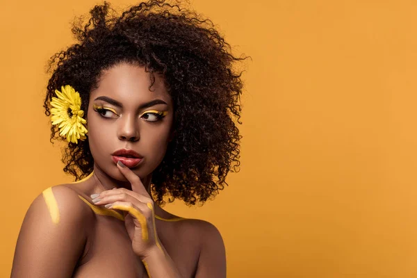 Joven mujer afroamericana brillante con maquillaje artístico y gerbera en el pelo sostiene la mano por su cara aislada sobre fondo naranja - foto de stock