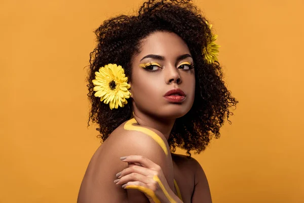 Joven mujer afroamericana sensual con maquillaje artístico y gerbera en el pelo mirando al lado aislado sobre fondo naranja - foto de stock