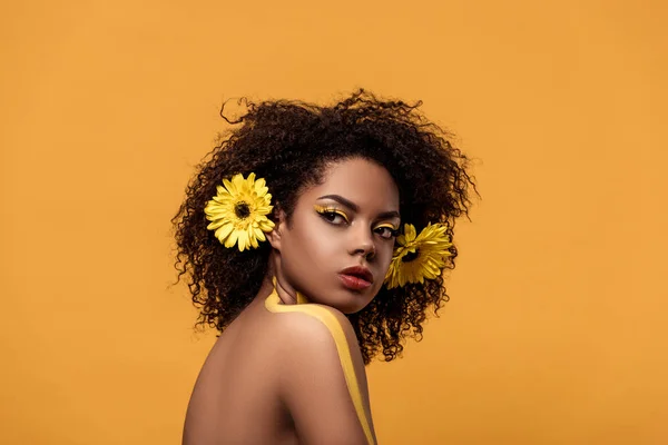 Joven mujer afroamericana brillante con maquillaje artístico y gerbera en cabello aislado sobre fondo naranja - foto de stock