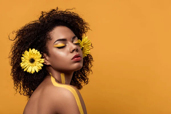 Joven mujer afroamericana brillante con maquillaje artístico y gerbera en el cabello soñando aislado sobre fondo naranja - foto de stock