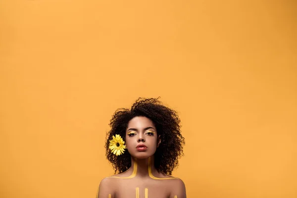 Joven mujer afroamericana sensual con maquillaje artístico y gerbera en el pelo mirando a la cámara aislada sobre fondo naranja - foto de stock