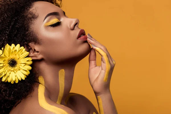Junge helle afrikanisch-amerikanische Frau mit artistischem Make-up und Gerbera im Haar mit der Hand am Kinn isoliert auf orangefarbenem Hintergrund — Stockfoto