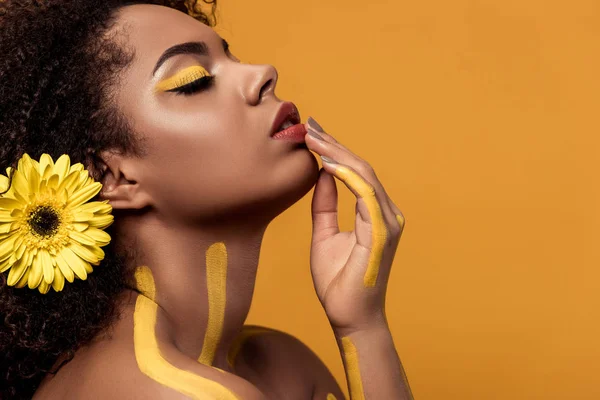 Young bright african american woman with artistic make-up and gerbera in hair with hand on chin isolated on orange background — Stock Photo