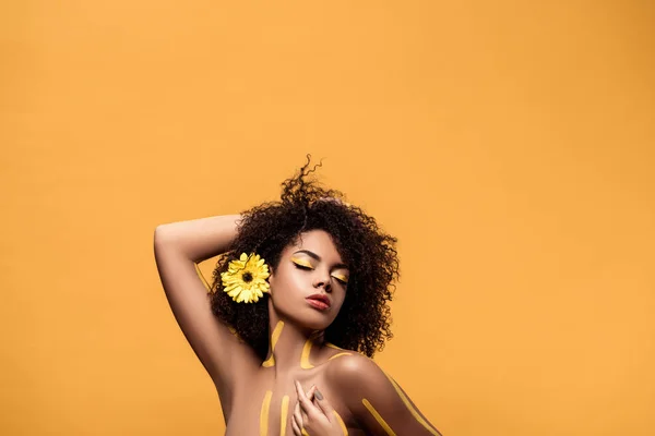 Joven mujer afroamericana sensual con maquillaje artístico y gerbera en el pelo, brazo detrás de su cabeza aislado sobre fondo naranja - foto de stock