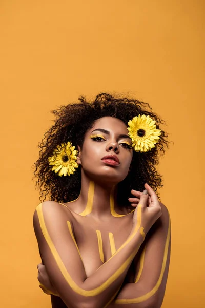 Joven mujer afroamericana con maquillaje artístico y gerbera en pelo de pie con brazos cruzados aislados sobre fondo naranja - foto de stock