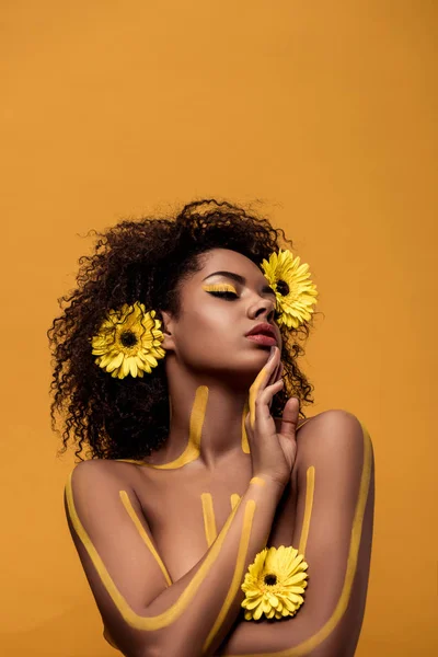 Молодая нежная африканская американка с художественным макияжем и герберой в волосах, изолированных на оранжевом фоне — стоковое фото