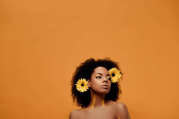 Joven mujer afroamericana sensual con maquillaje artístico y gerbera en pelo aislado sobre fondo naranja - foto de stock