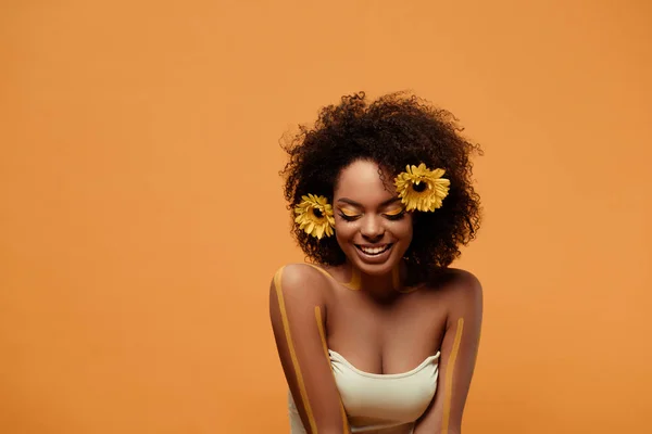 Jeune femme afro-américaine riante avec maquillage artistique et gerbera dans les cheveux isolés sur fond orange — Photo de stock