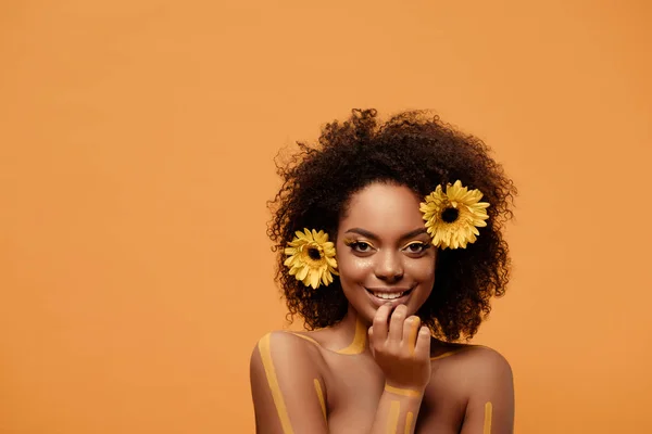 Giovane donna afroamericana sorridente con trucco artistico e gerbera in capelli isolati su sfondo arancione — Foto stock