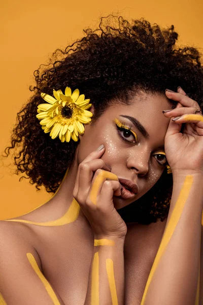 Beautiful african american woman with artistic make-up and gerbera in hair with hands near face isolated on orange background — Stock Photo