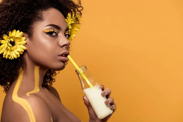 Atractiva joven afroamericana con maquillaje artístico y gerberas en el cabello bebiendo leche de botella aislada sobre fondo naranja - foto de stock