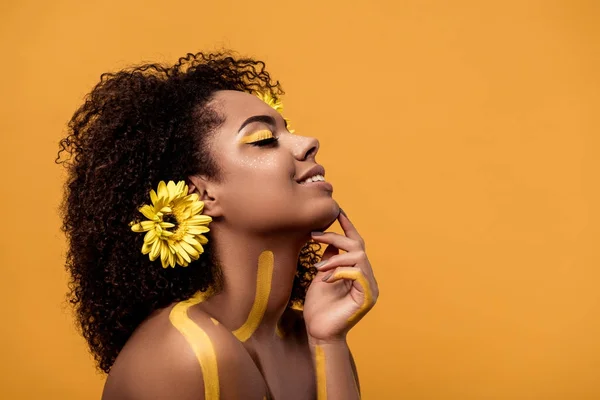 Jeune femme afro-américaine tendre avec maquillage artistique et gerbera dans les cheveux isolés sur fond orange — Photo de stock