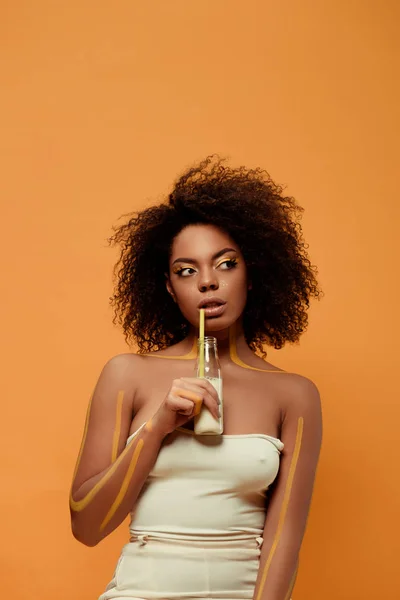 Femme afro-américaine élégante avec maquillage artistique boire du lait de bouteille isolé sur fond orange — Photo de stock