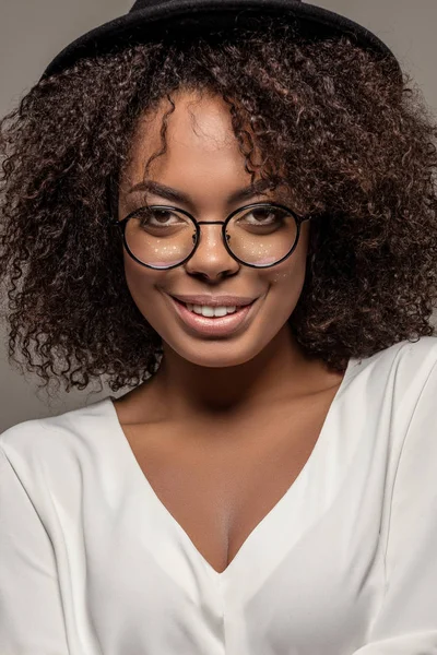 Young smiling african american woman in white shirt wearing glasses and hat isolated on grey background — Stock Photo