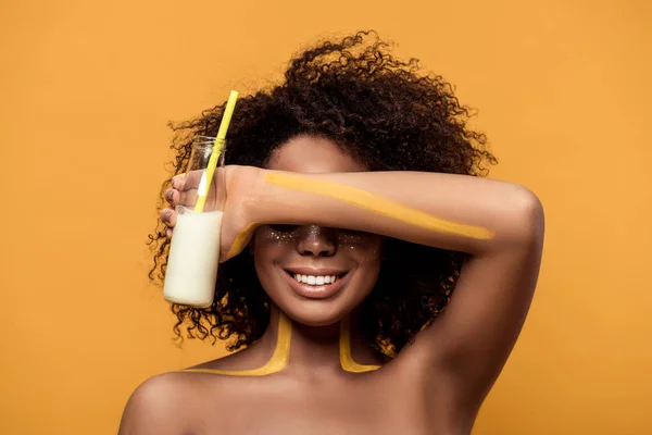 Joven mujer afroamericana sonriente con maquillaje artístico sosteniendo botella con leche aislada sobre fondo naranja - foto de stock