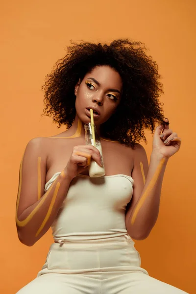 Joven mujer afroamericana sensual con maquillaje artístico bebiendo leche de botella aislada sobre fondo naranja - foto de stock
