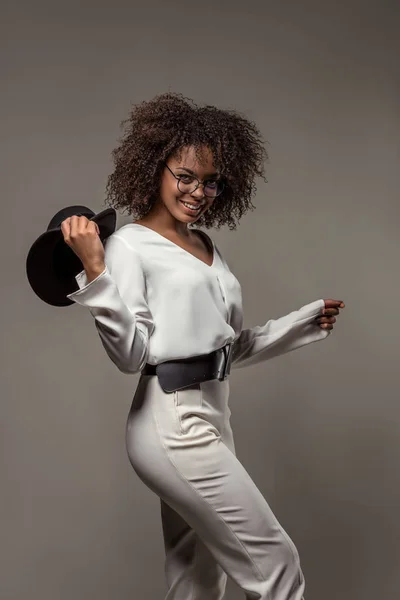 Стильная африканская американка в белой одежде улыбается и машет черной шляпой на сером фоне — стоковое фото