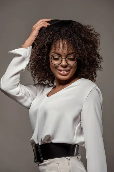 Young sensual african american woman in white shirt wearing glasses holds hat on her head isolated on grey background — Stock Photo