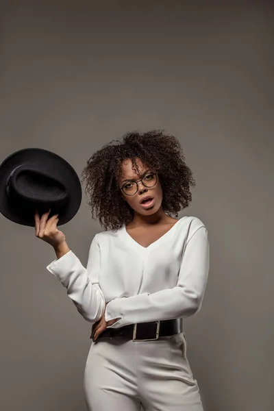 Young angry african american woman in white shirt wearing glasses holds hat isolated on grey background — Stock Photo