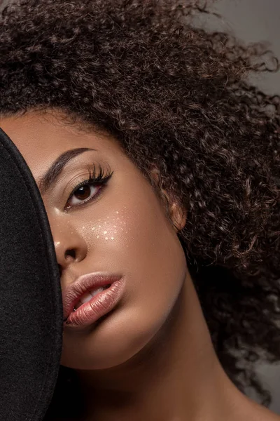 Young sensual african american woman holding black hat over half of her face isolated on grey background — Stock Photo