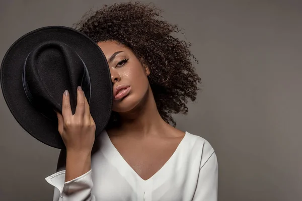 Atraente jovem afro-americana de camisa branca segurando chapéu preto sobre metade de seu rosto isolado em fundo cinza — Fotografia de Stock